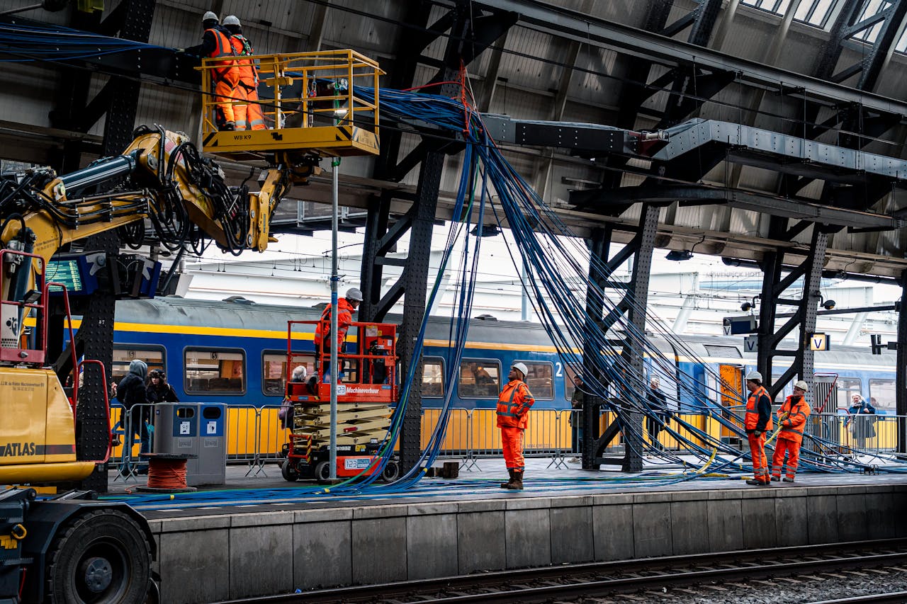 Team of workers in safety gear installing cables at a busy train station platform.